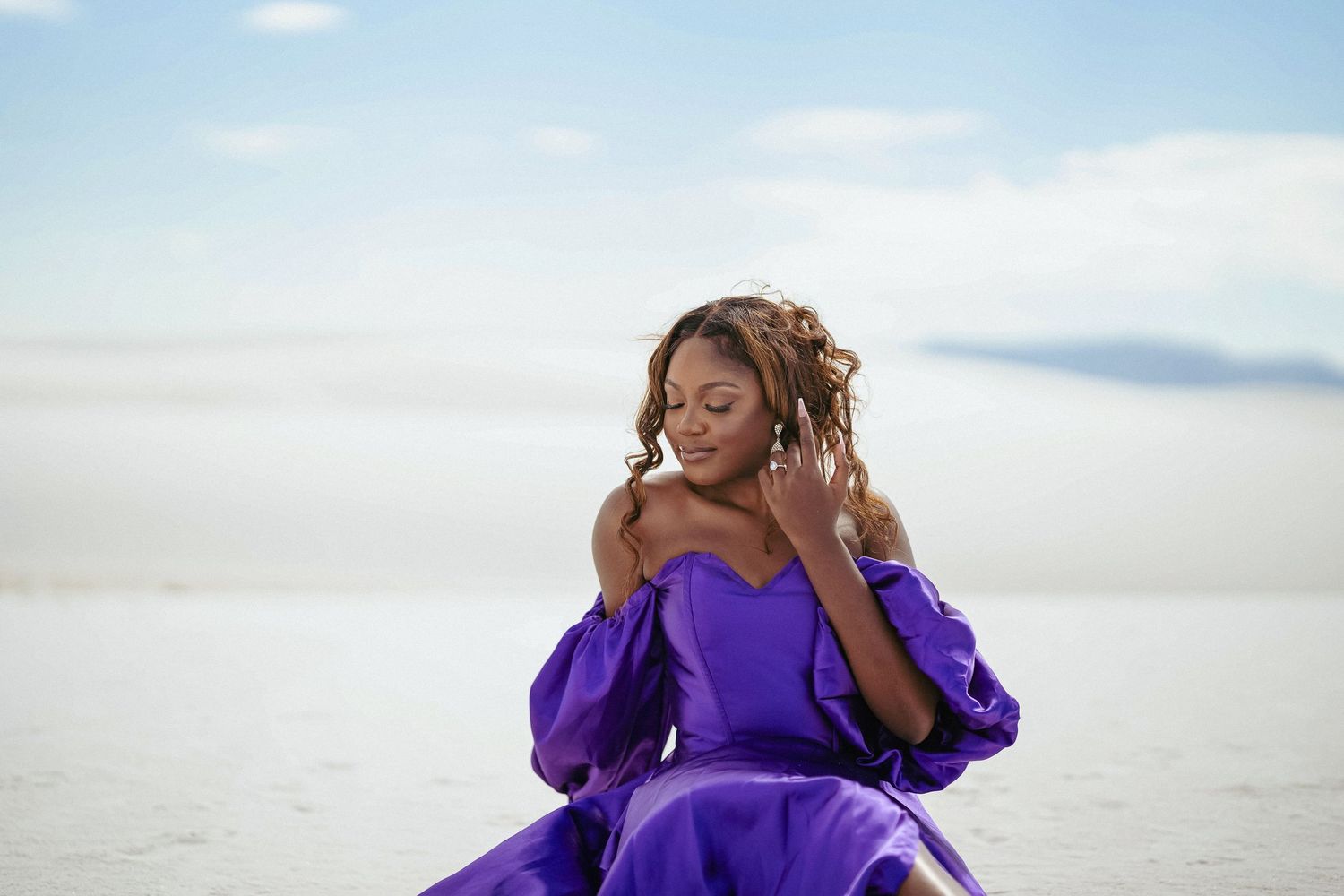 A person in purple dress poses playfully on white sand against cloudy sky.