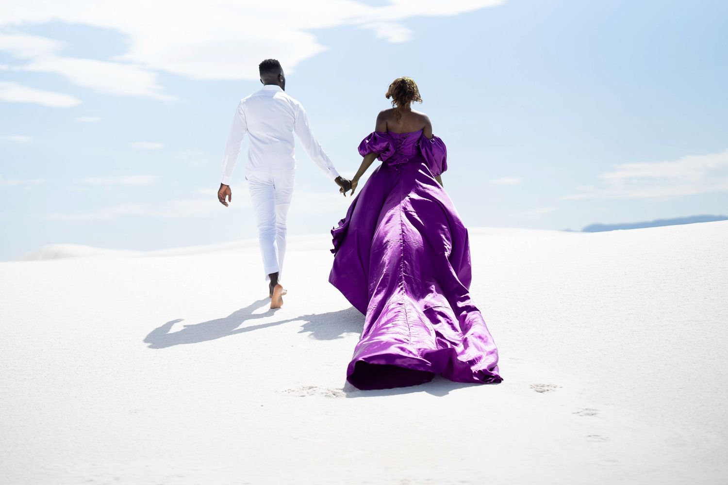 Two figures in contrasting purple and white outfits walk across vast white sand dunes.