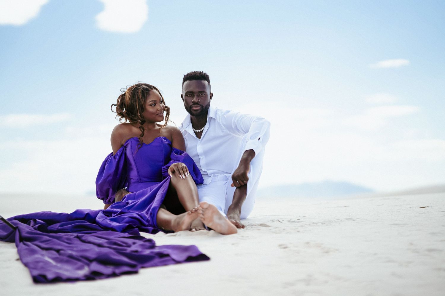 Loving couple relaxes together on pristine white sand beach with purple dress flowing in breeze.