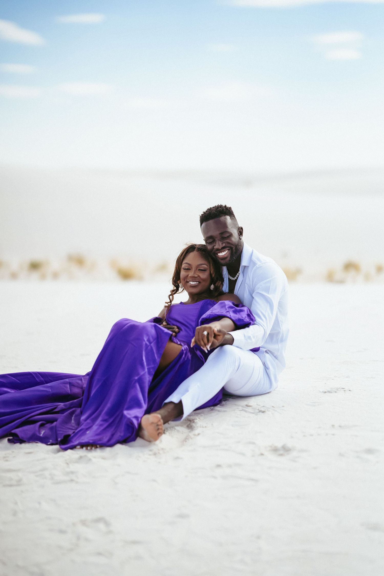 Couple shares tender moment while lying on sandy beach with purple dress billowing.