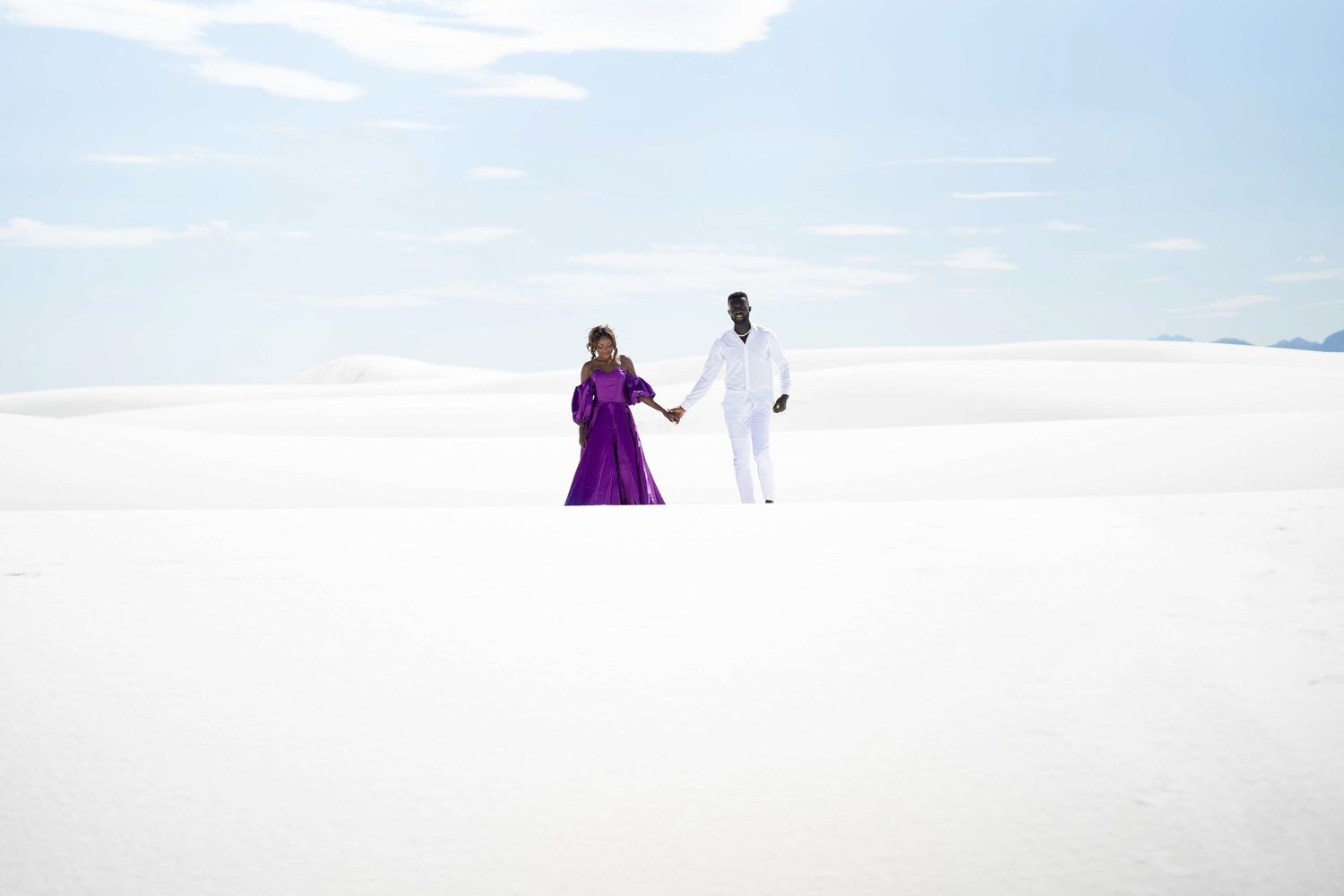 Distant figures in purple and white create striking silhouettes against white sand dunes.