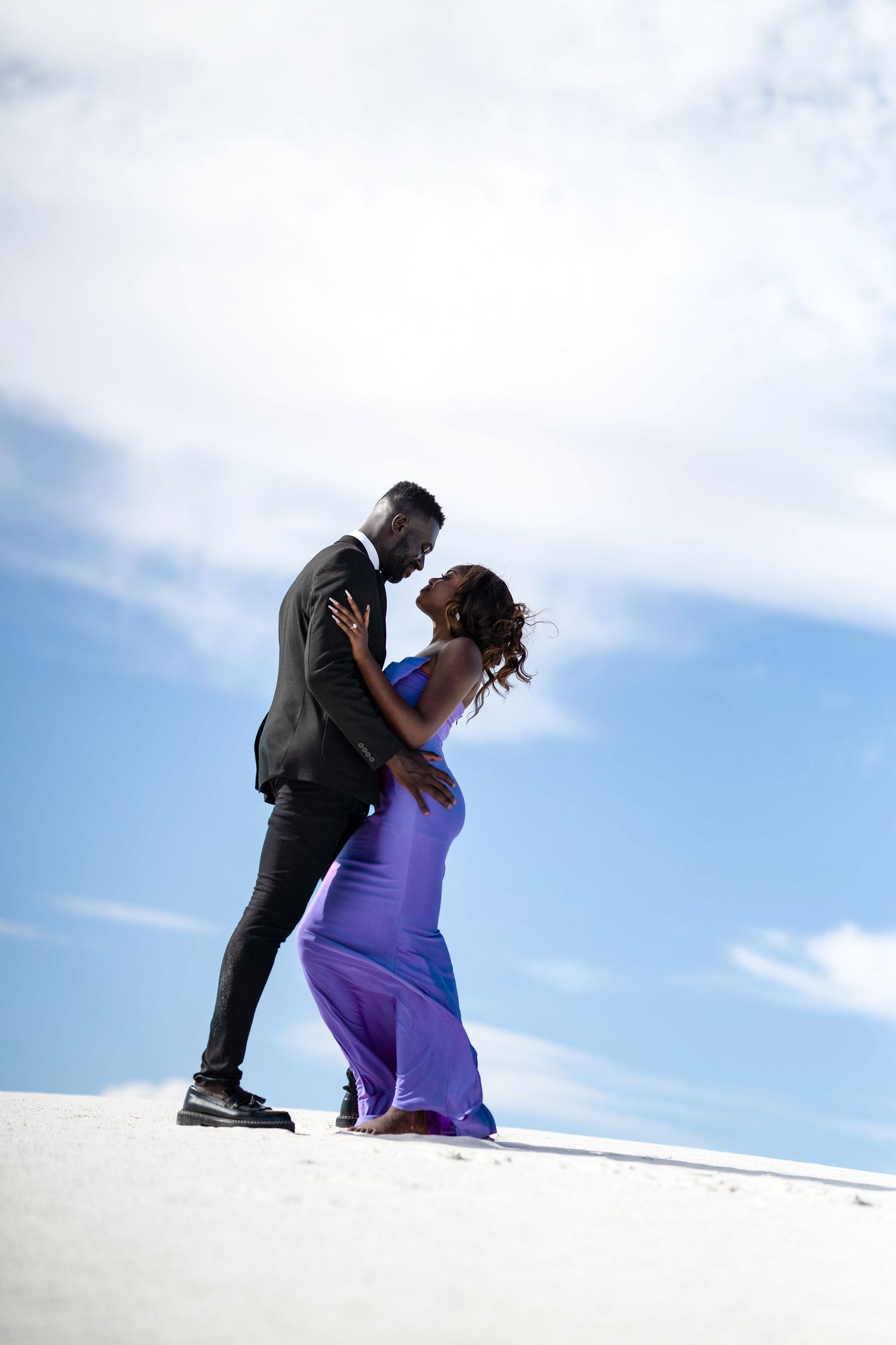 Couple stands atop sandy dune with purple dress flowing dramatically in wind.
