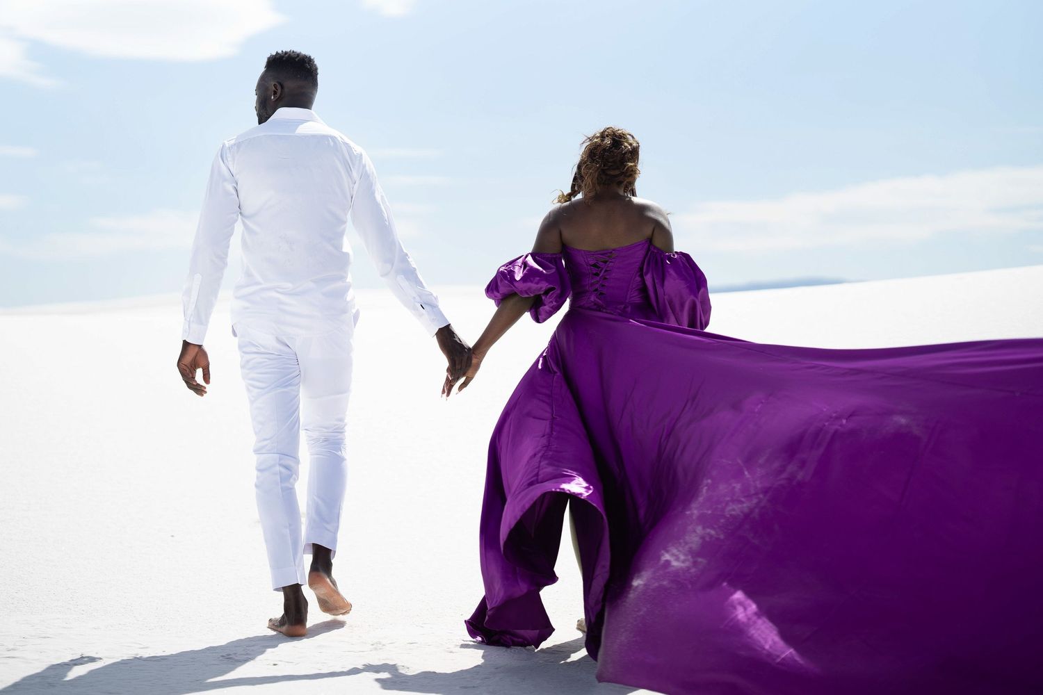 Couple walks along beach with flowing purple dress creating dramatic silhouette against sky.