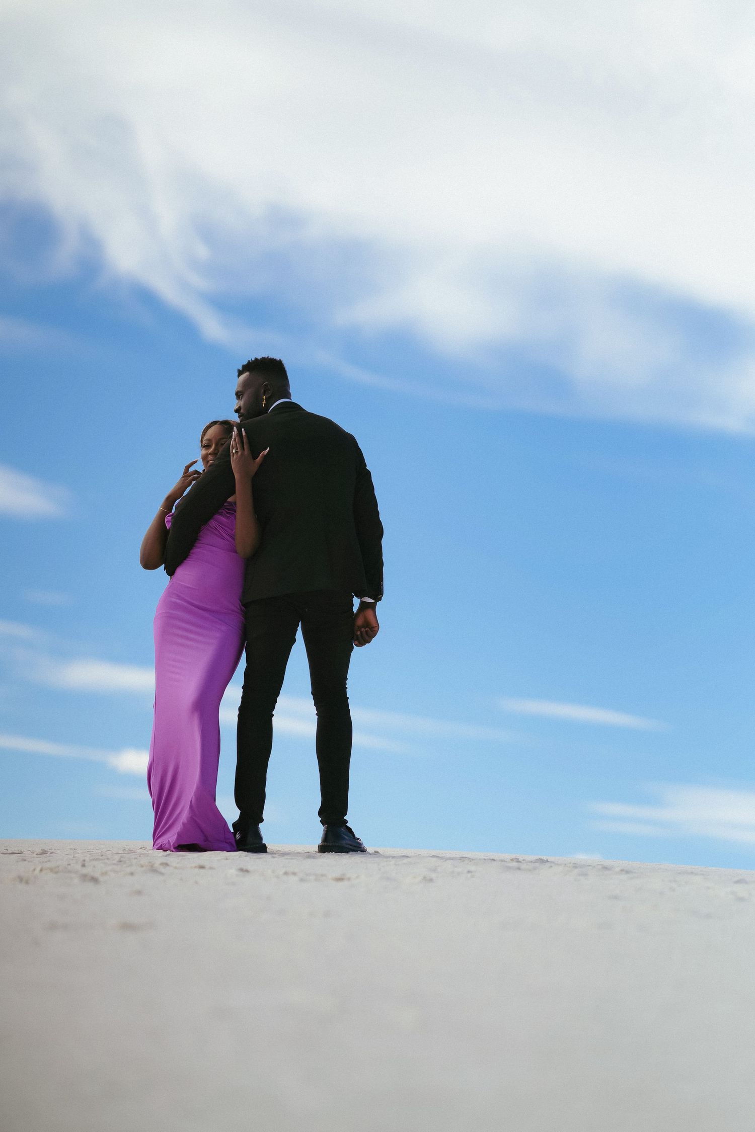 Two figures embrace on hilltop with purple dress contrasting against blue sky.
