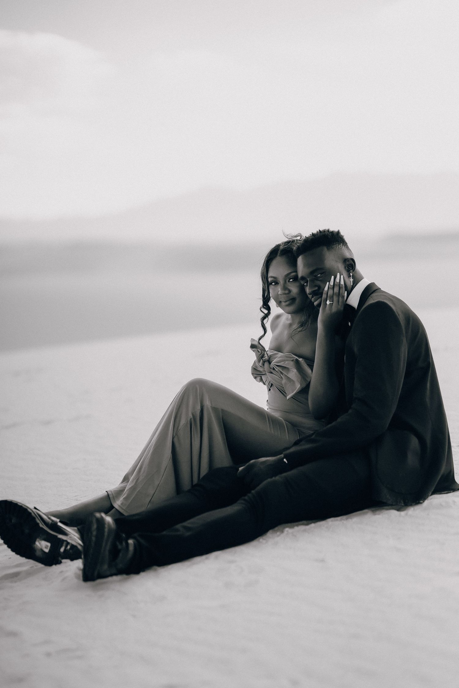A romantic black and white photo of a couple sitting together on white sand with mountains in the distance.