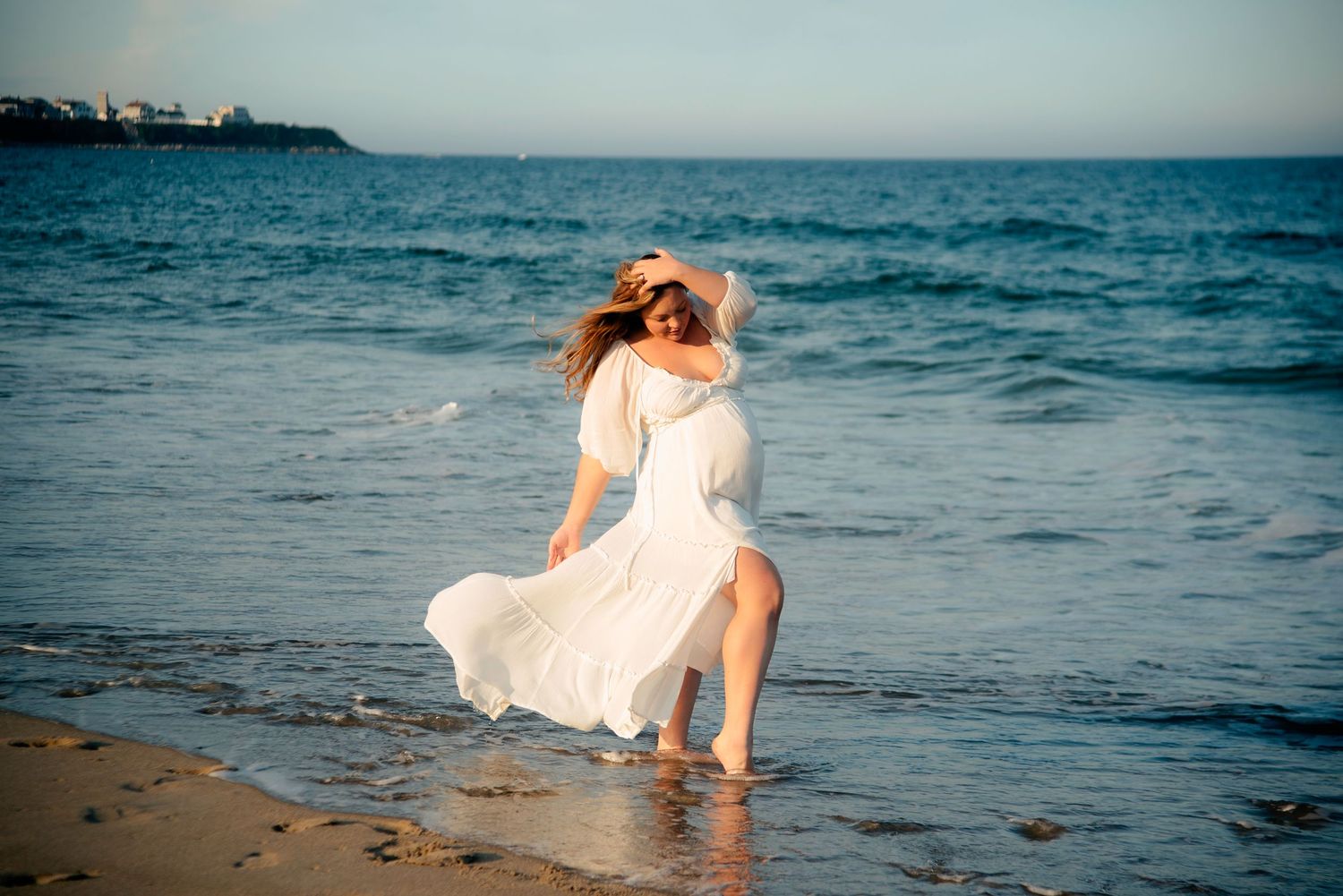 pregnant mama posing in a flowing dress while the wind sweeps  her dress behind her in an outdoor beach maternity photoshoot.