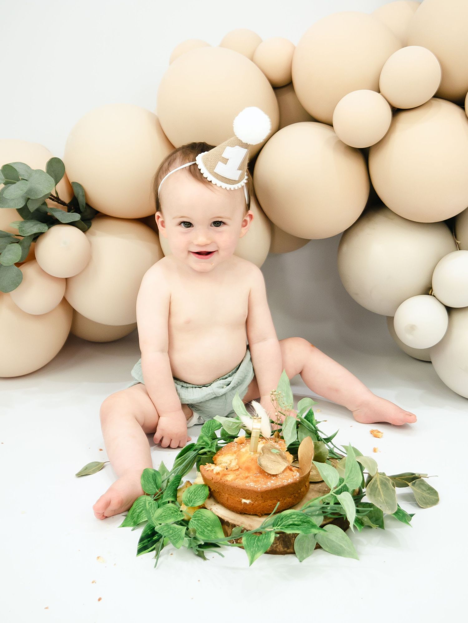 Whimsical cake smash photo setup with neutral balloon arch, eucalyptus leaves, and small dessert on white background.