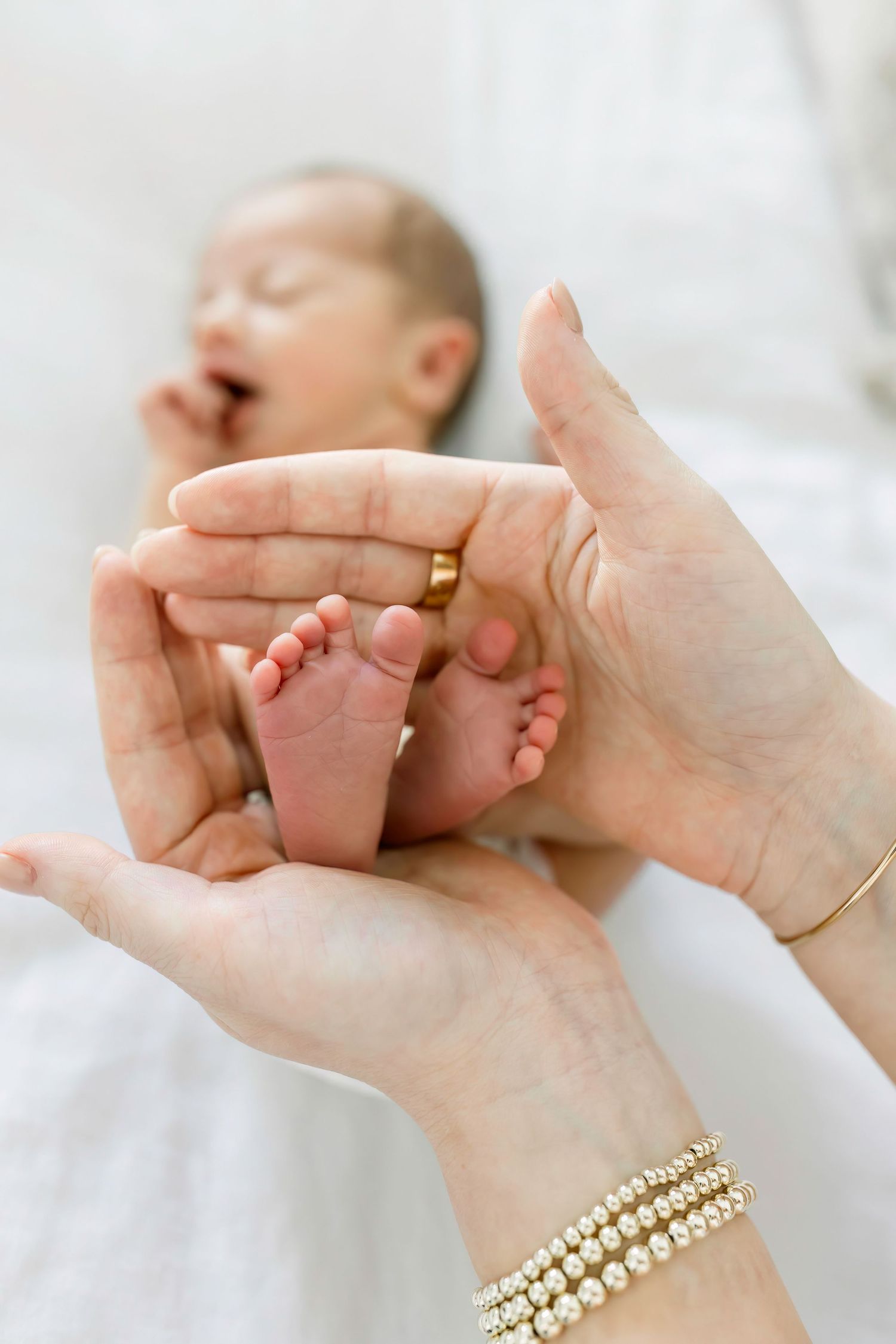 Tiny newborn baby feet cradled in adult hands wearing gold jewelry and bracelets on white bedding.