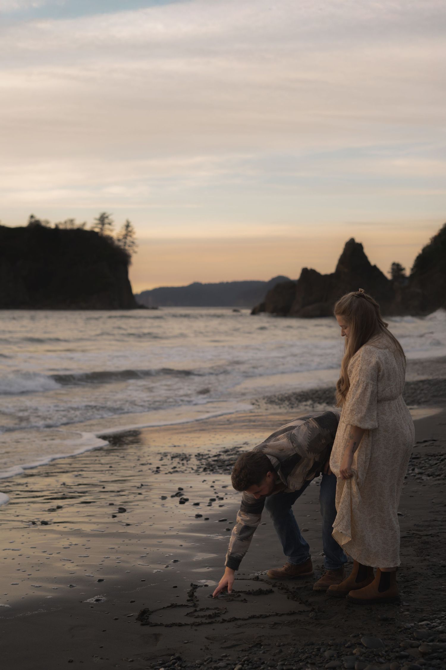 People writing in sand on a dramatic rocky beach at sunset.