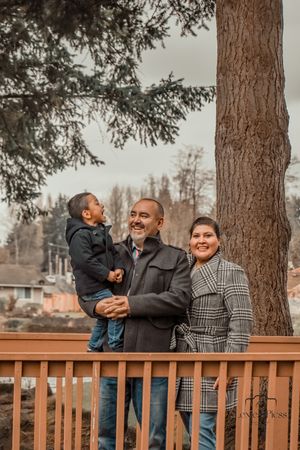 Family poses together on a wooden deck against a backdrop of tall evergreen trees.