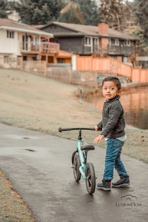 Small child plays with light blue balance bike on a suburban sidewalk with houses in the background.