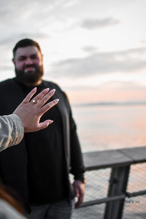 Hand displaying engagement ring with person in black sweater against sunset backdrop.