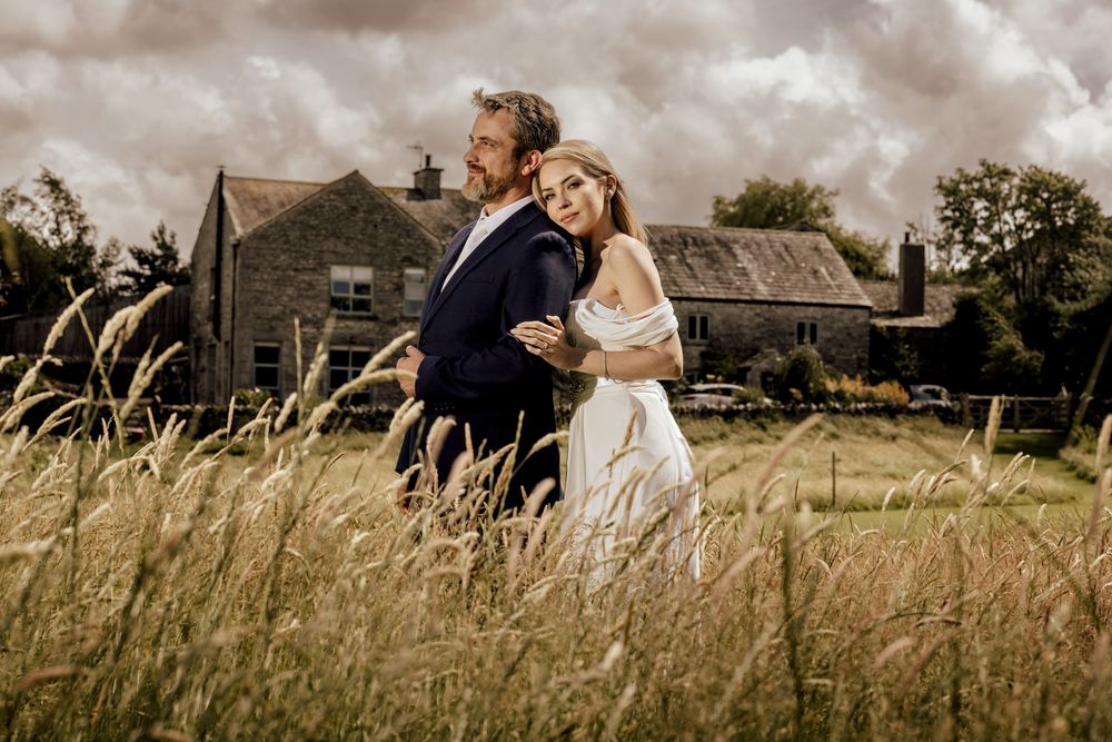 A bride and groom embrace in a golden wheat field with a rustic stone farmhouse in the background under dramatic cloudy skies.