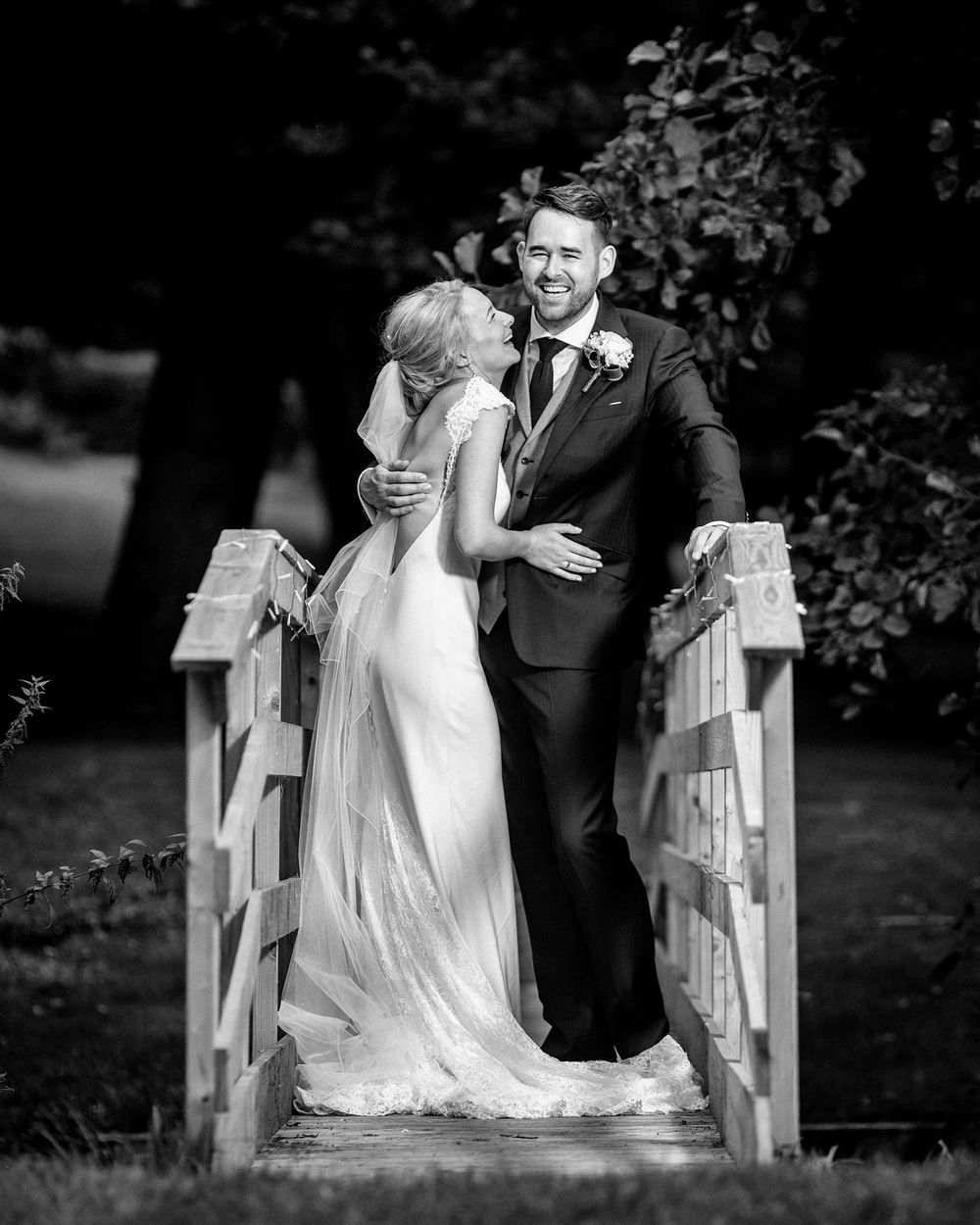 Black and white photo of a bride and groom sharing a romantic moment on a wooden bridge surrounded by lush greenery.