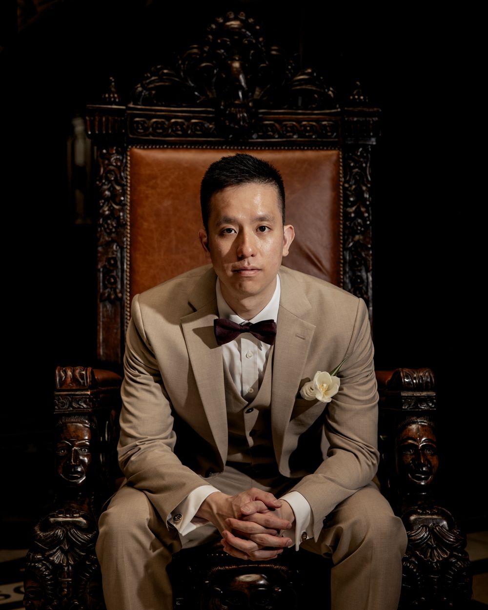 A groom in a tan suit with bow tie sits regally in an ornate wooden throne chair, hands clasped, with a white boutonniere.