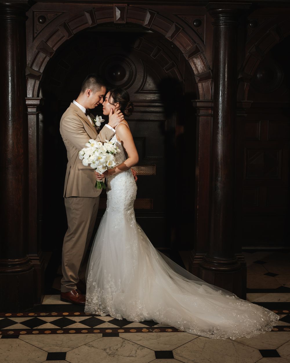 Bride and groom share romantic moment in mermaid lace gown and tan suit, holding white bouquet in ornate church doorway.