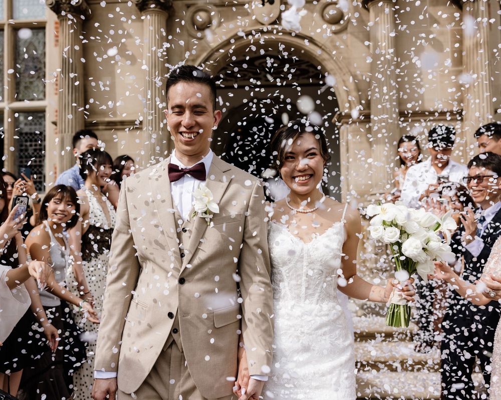 Newlywed couple smiling as confetti falls outside a church, bride holding white bouquet in elegant wedding dress.