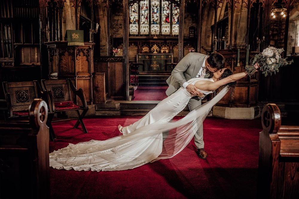 Bride and groom share a romantic dip kiss inside a stunning church with red carpet and stained glass windows.