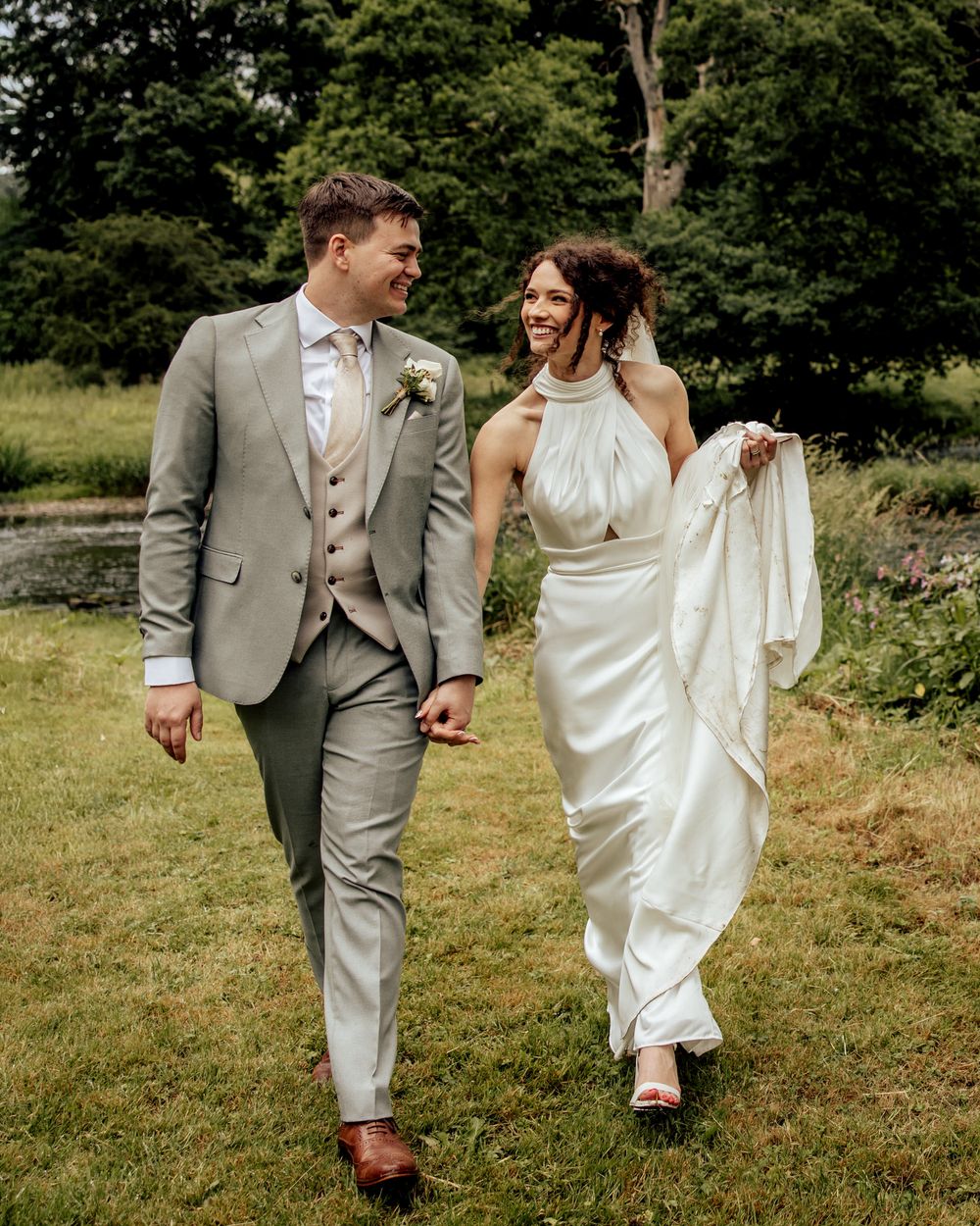 A smiling bride in a white halter gown and groom in a sage suit walk hand in hand through a lush green garden.