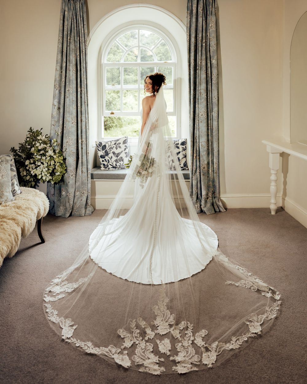 Bride in elegant white wedding gown with long lace-trimmed cathedral veil standing by arched window in ornate room.