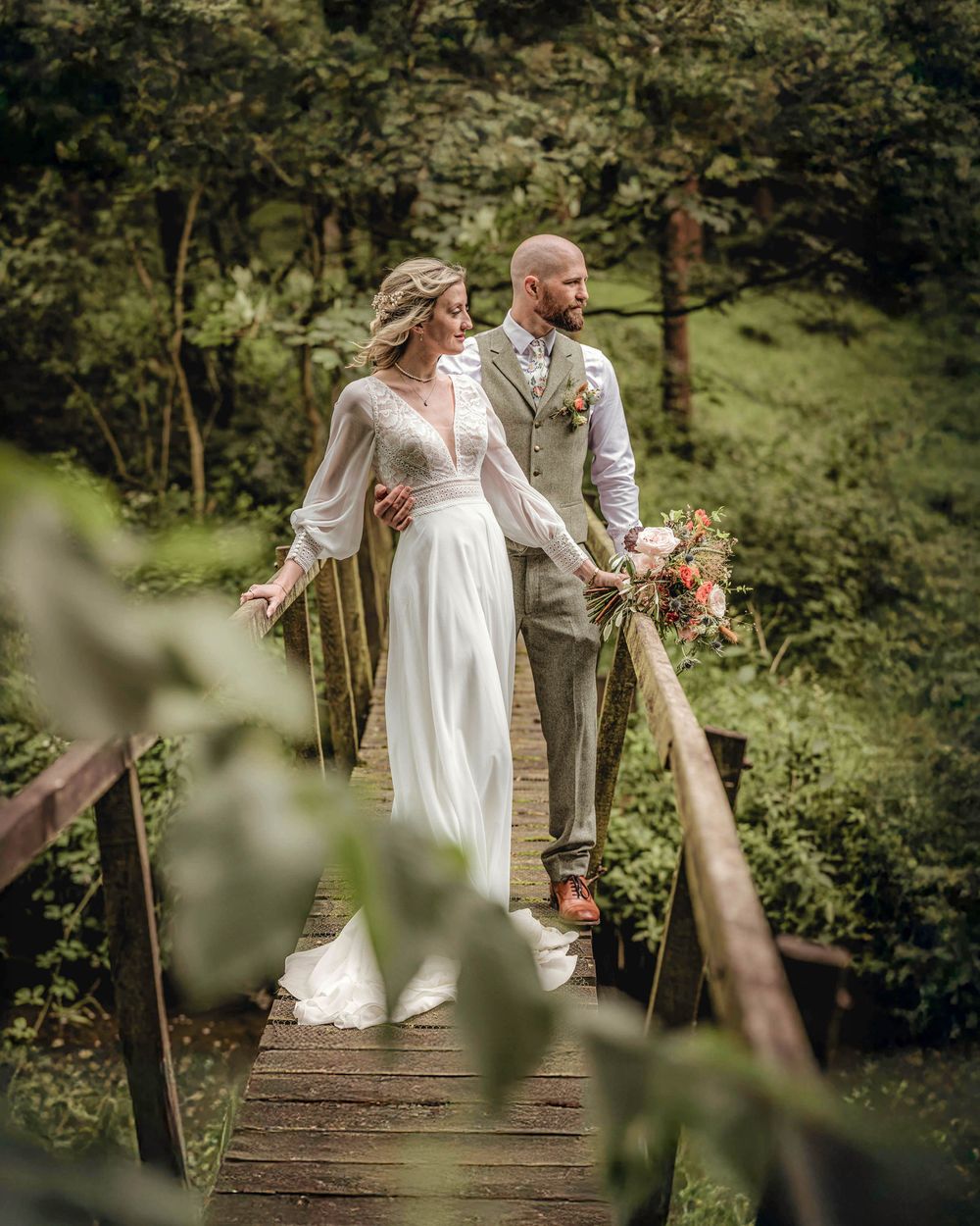 Bride and groom walking on a wooden bridge surrounded by lush greenery, bride holding a floral bouquet in a white gown.
