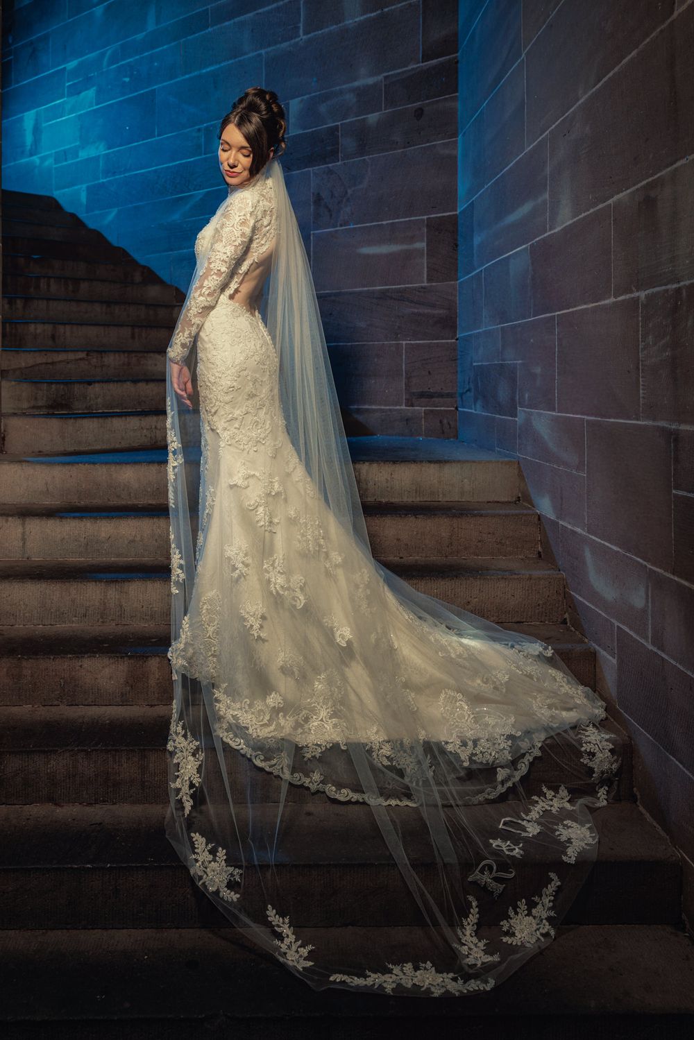 Bride in lace mermaid wedding gown with long veil stands on stone staircase, smiling elegantly.