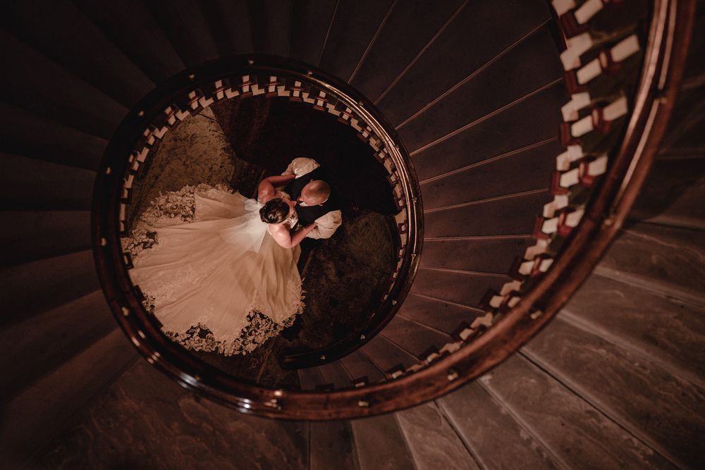Overhead view of a bride in a lace wedding gown and a groom embracing at the center of a elegant wooden spiral staircase.