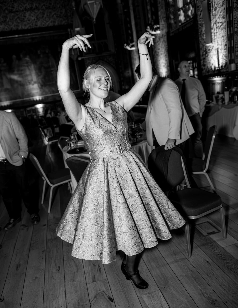 Woman in a brocade full-skirted dress dances joyfully with arms raised at a wedding reception in a wood-floored hall.