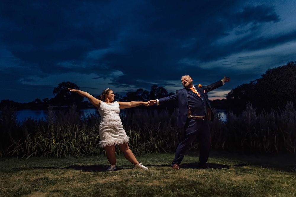 Couple in wedding attire playfully lean back holding hands outdoors under dramatic evening sky.