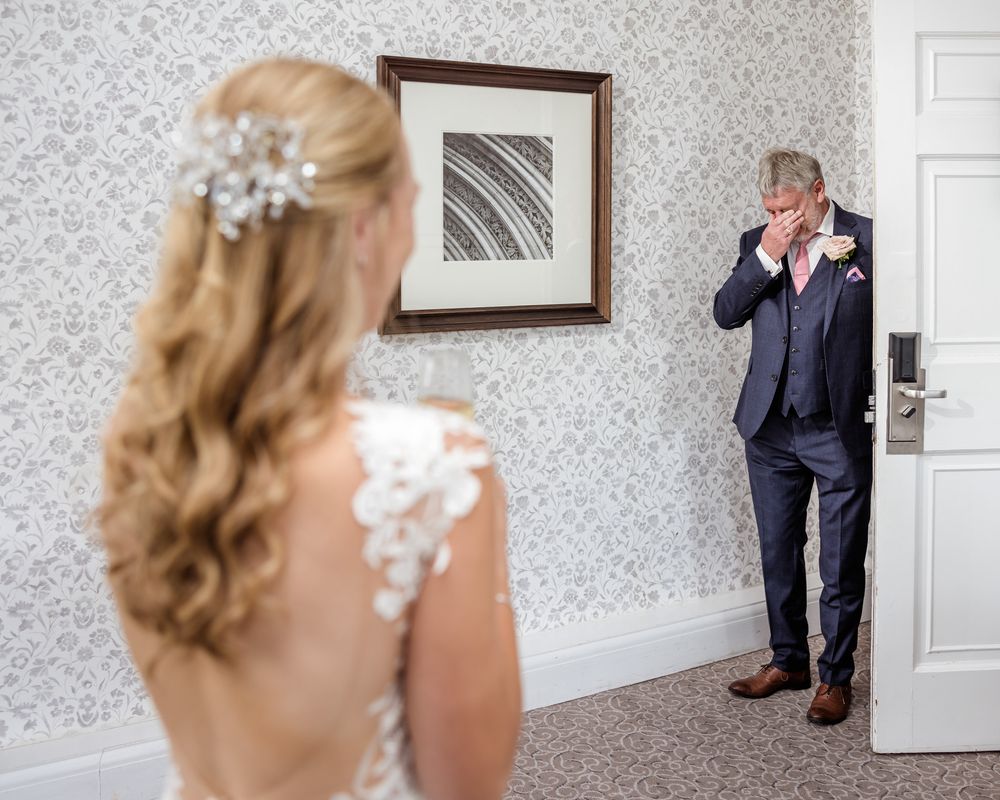 An emotional first look moment between a bride in a lace gown and her father in a navy suit at a wedding venue.