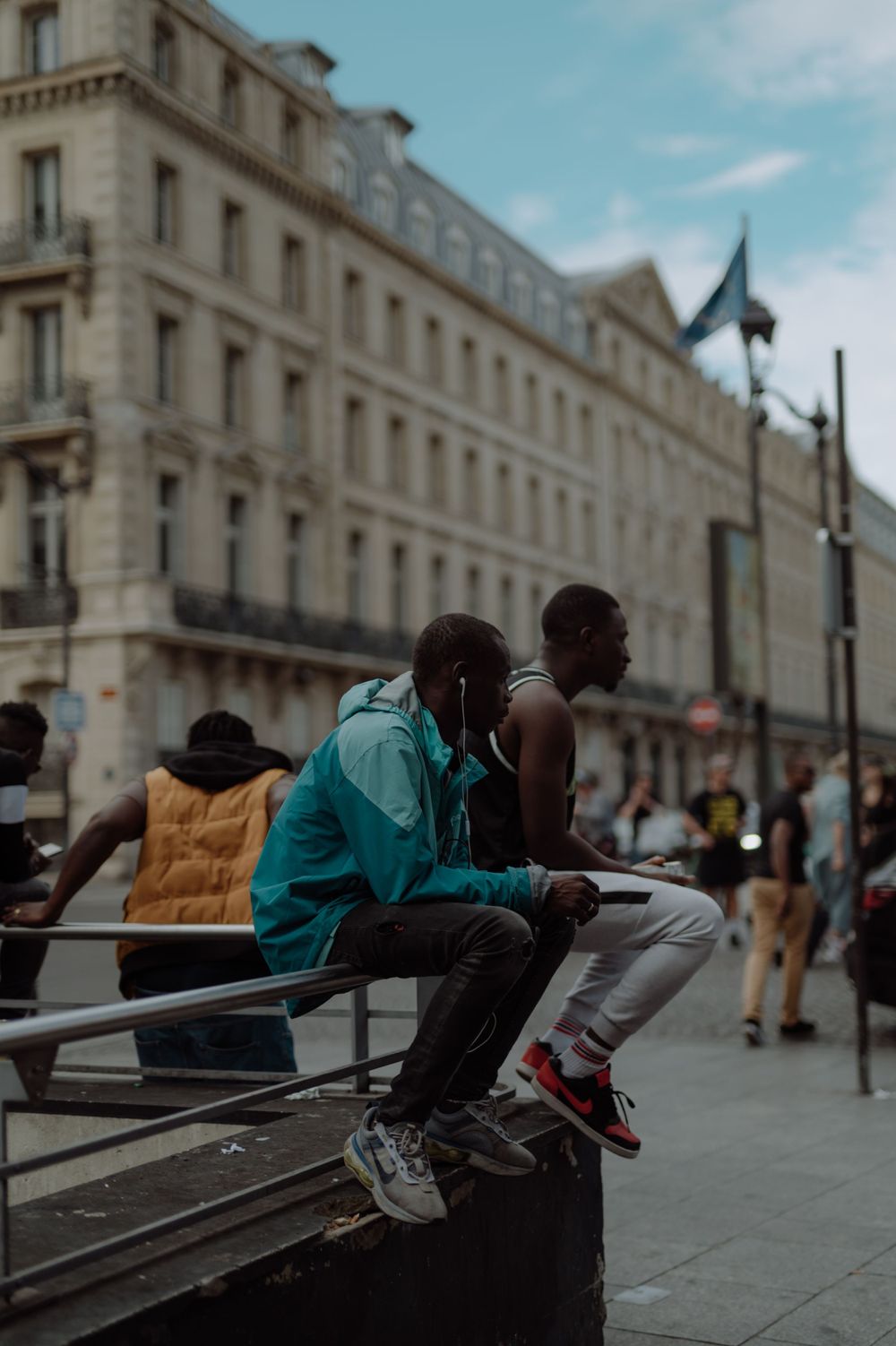 Two friends sit casually on metal railings against a backdrop of classic Parisian architecture.