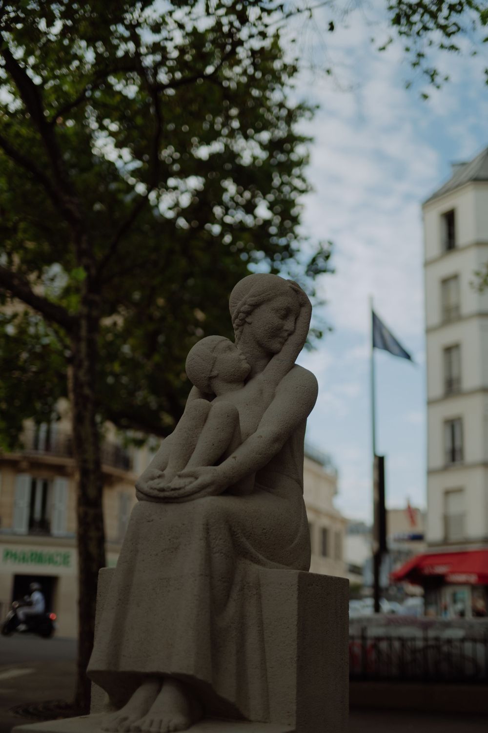 Stone sculpture of a mother and child sitting together in front of Parisian buildings.