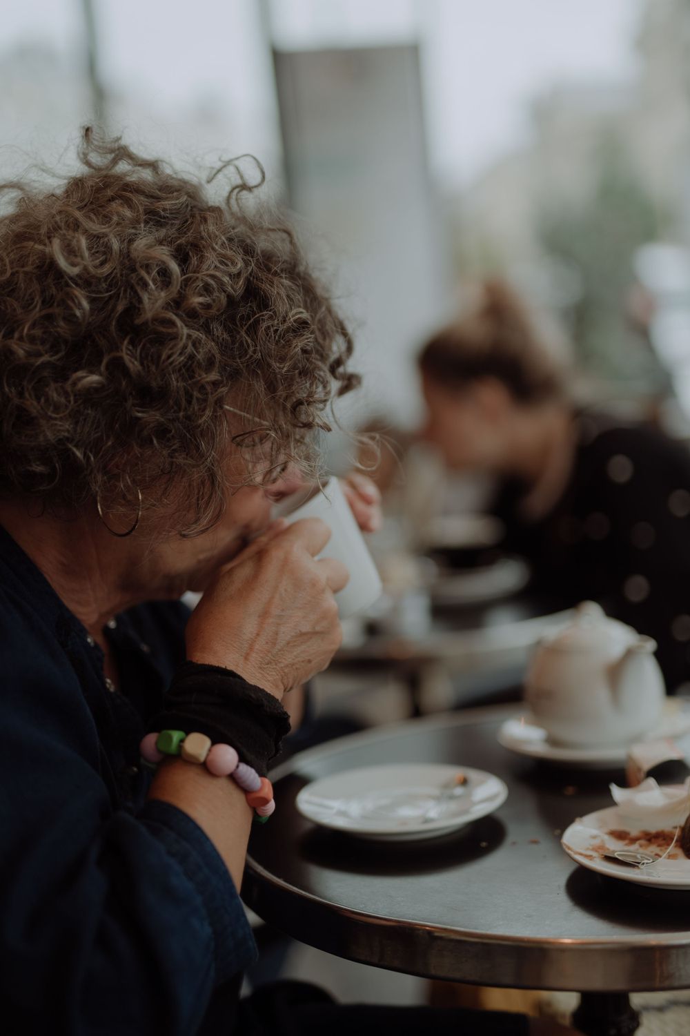 Person with curly hair drinking from a cup at a cafe table in moody lighting.