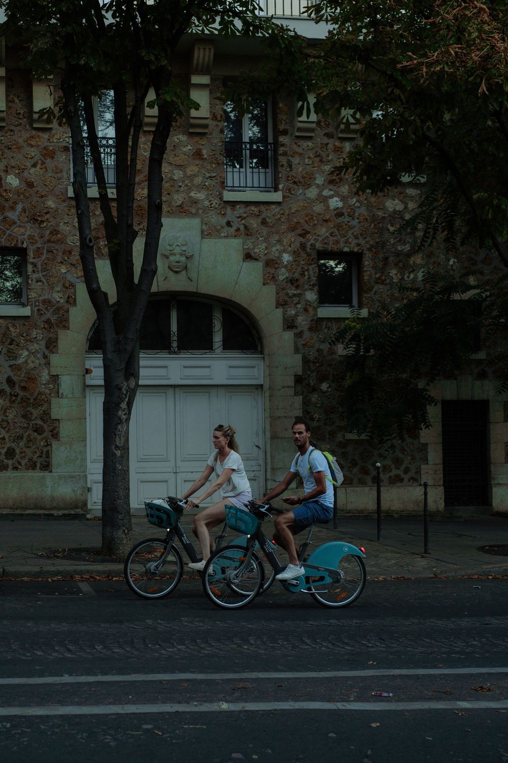 Two cyclists riding blue rental bikes past a historic stone building.