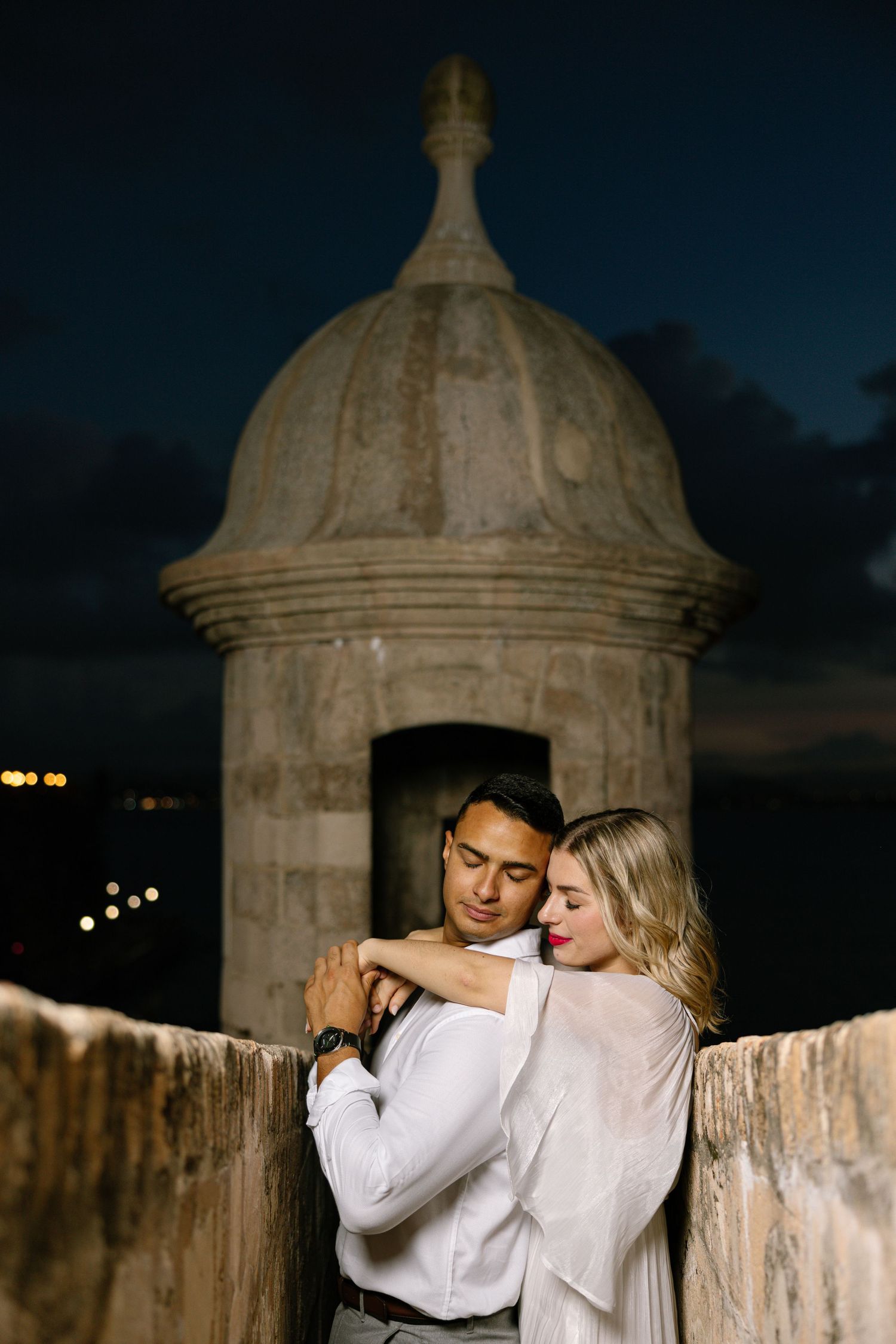 Romantic couple embraces at night by historic stone turret with domed architecture against dark sky.
