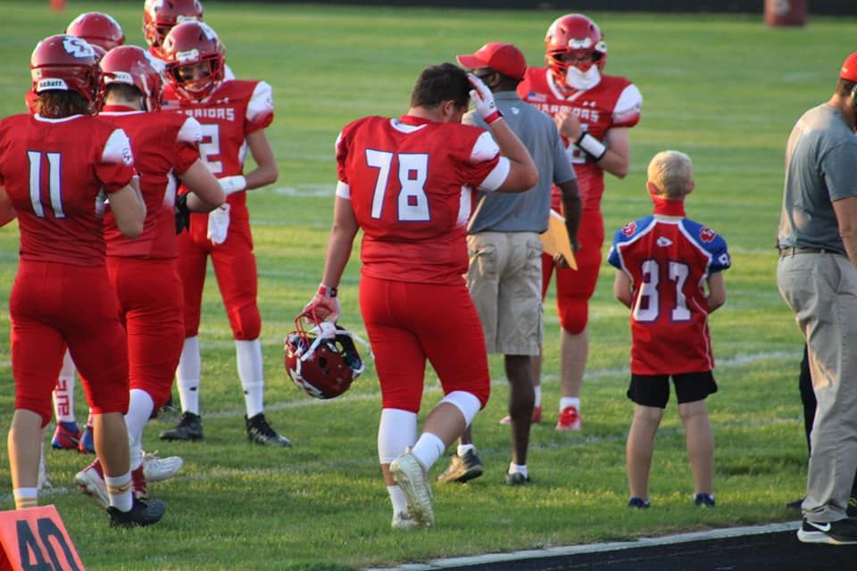 Red football team players in uniform on field, player 78 holding helmet, with coaches and young child nearby.