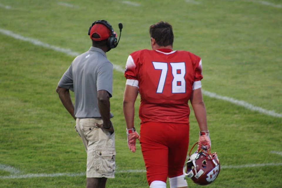 Football player #78 in red jersey walks with coach wearing headset on green field sideline.