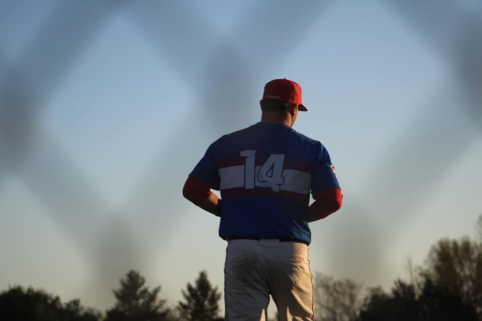 Baseball player wearing number 14 blue jersey and red cap stands near chain-link fence during golden hour.