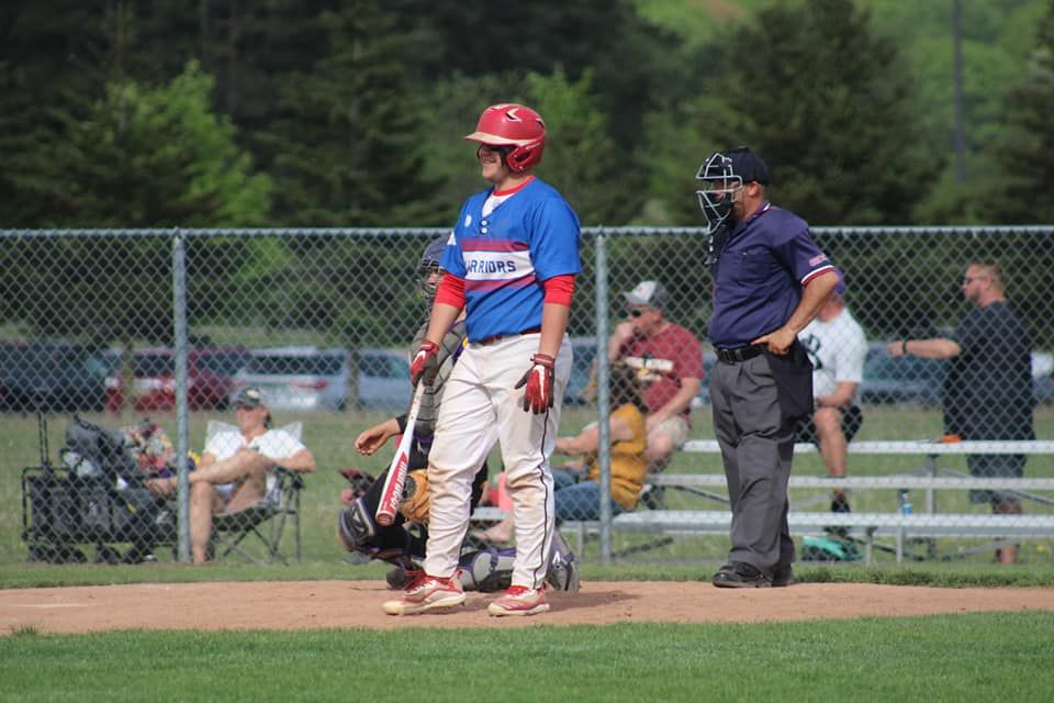 Baseball batter in red and blue uniform stands at home plate with umpire and catcher during a youth baseball game.