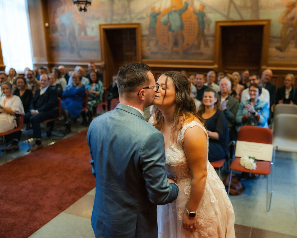 A romantic wedding ceremony kiss between newlyweds in a historic building with painted murals and guests looking on.