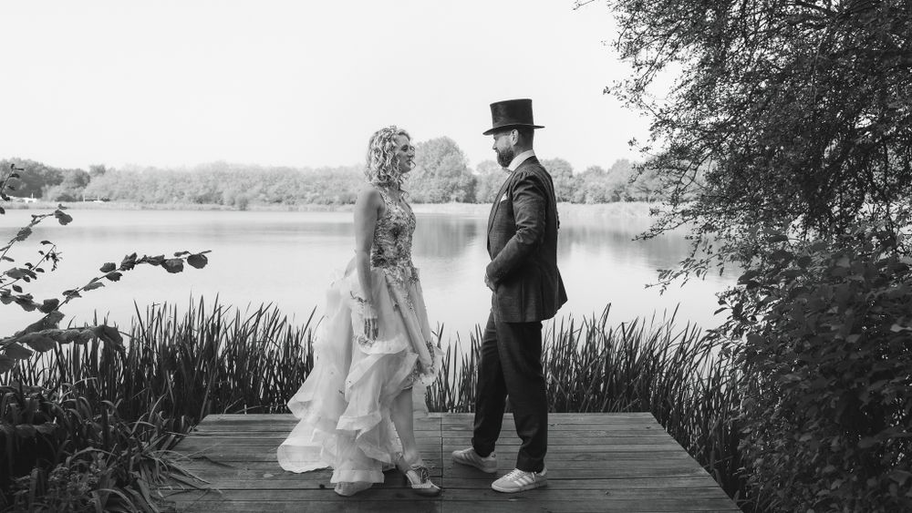 A black and white wedding photo of a couple standing on a wooden dock by a serene lake surrounded by nature.

- BU Photography -