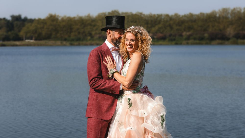 A romantic couple in wedding attire embraces by a serene lake with autumn trees in the background.

- BU Photography -