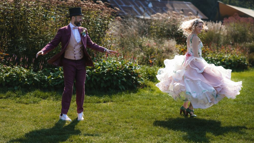Two people dance joyfully on a sunny lawn wearing formal attire including a pink flowing dress and brown suit.

- BU Photography -