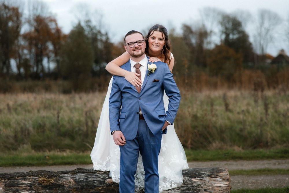 A couple in wedding attire stands together outdoors in autumn, with the groom wearing a blue suit against a natural backdrop.