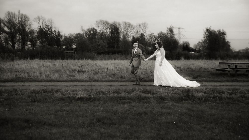 A black and white photo shows two figures walking across a grassy field, with one wearing a flowing white dress.