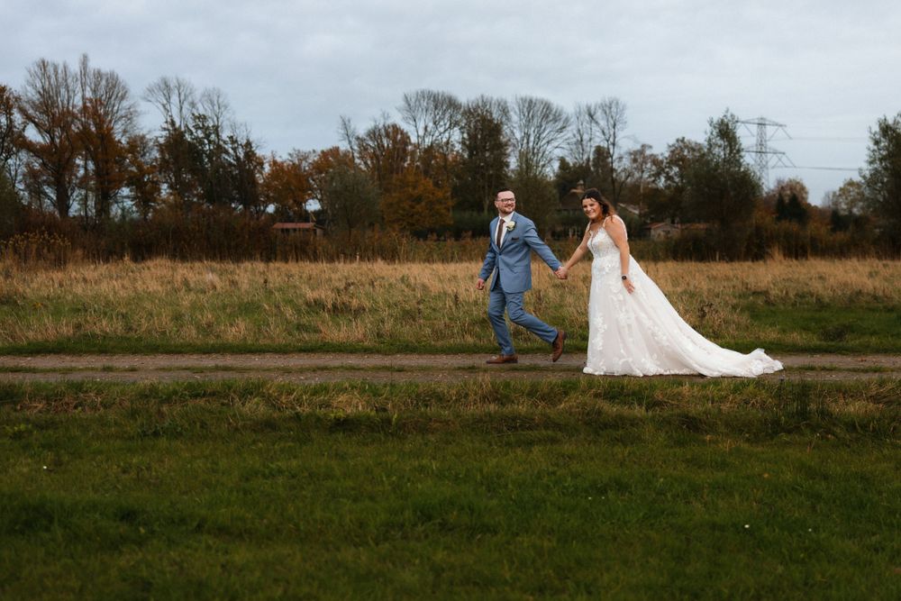 A couple in wedding attire runs playfully through an autumn field with bare trees in the background.