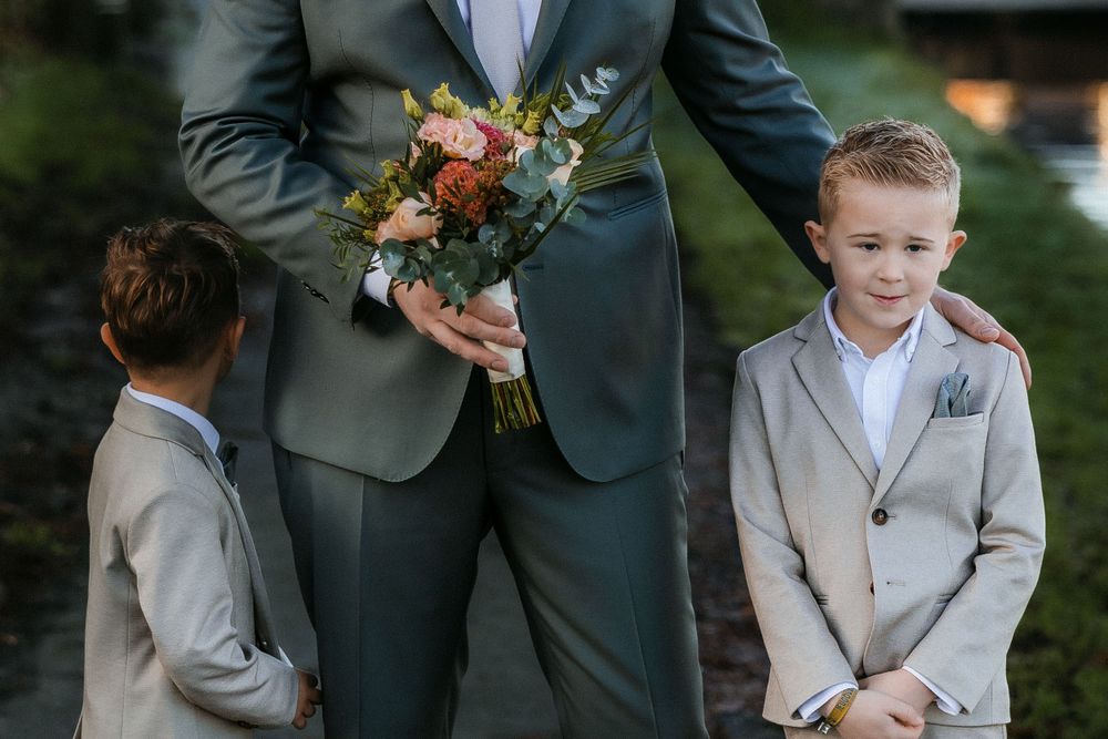 Small children in formal attire stand next to someone holding a wedding bouquet of peach and white flowers.
