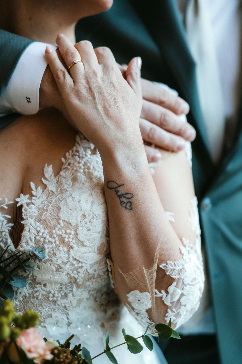 Close-up view of bride's hand with small wrist tattoo against white lace wedding dress and groom's suit.