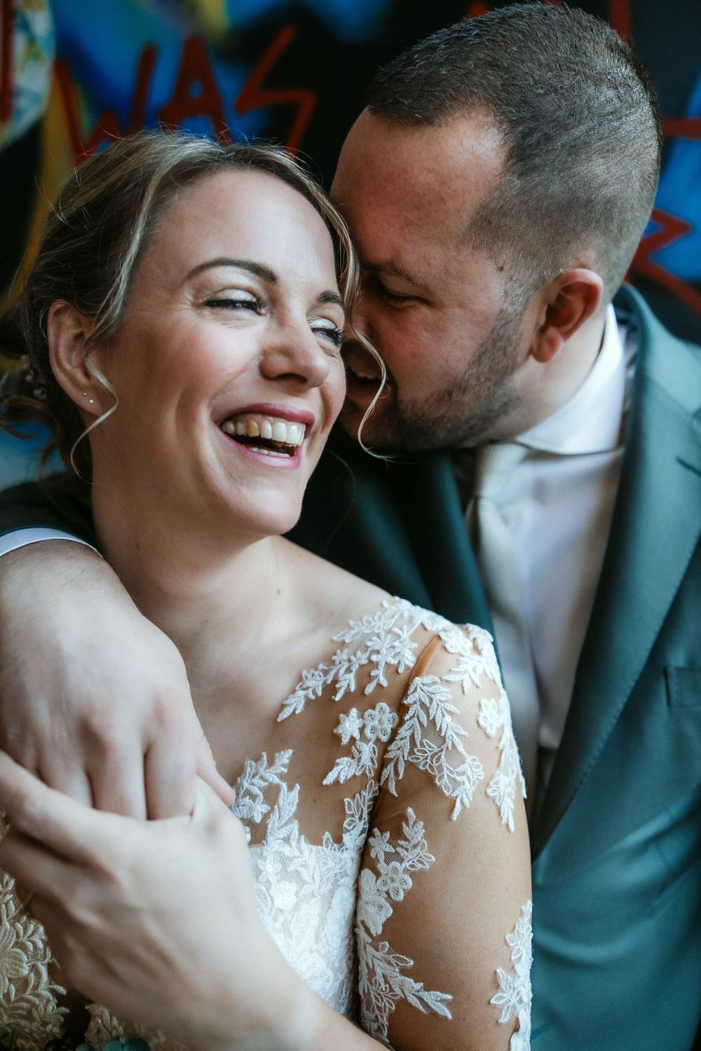 Bride in delicate lace sleeve dress laughs joyfully while groom leans in close against colorful graffiti wall background.