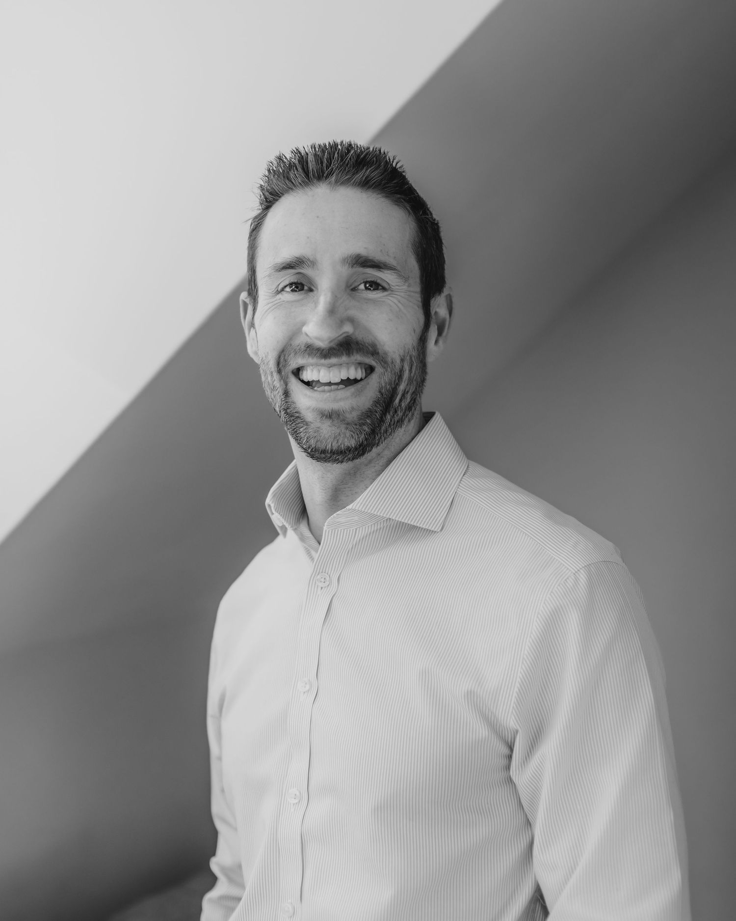 Professional headshot of a person in a white button-down shirt smiling against a gray background.